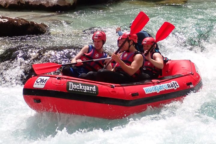 Four people in a small orange fun-raft with a guide on the turquoise Soča River, navigating a mild rapid, surrounded by green mountains.