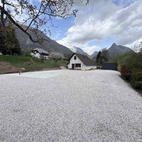 House with gravel yard, surrounded by mountains and trees under a cloudy sky.